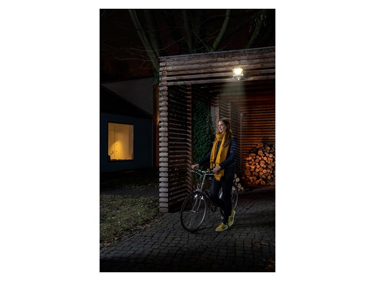 Frau mit Fahrrad unter einer Außenlampe an einem Holzschuppen mit Brennholz.