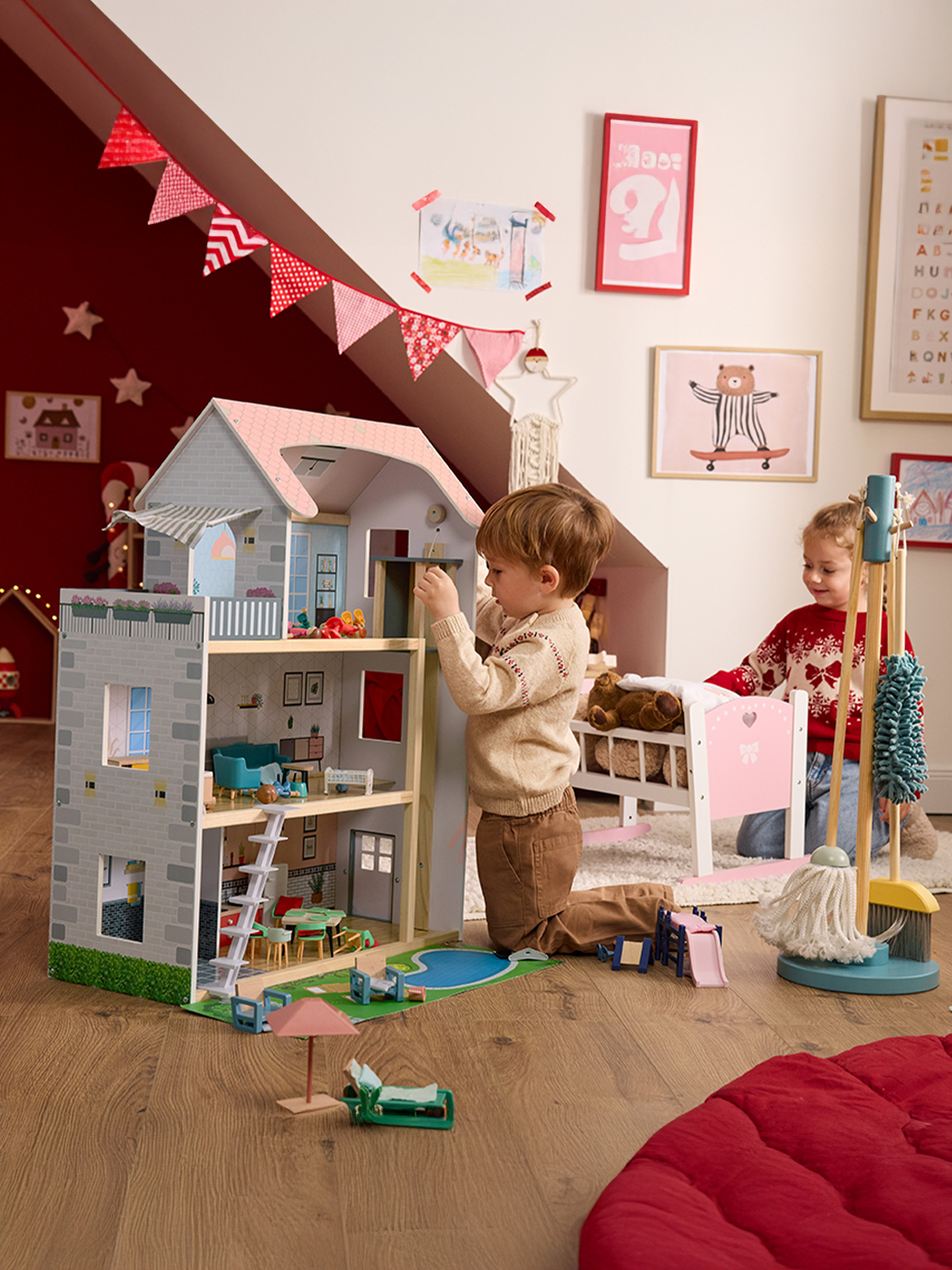 Zwei Kinder spielen mit einem großen Holzpuppenhaus und einem Spielzeug-Reinigungsset in einem festlichen Zimmer.