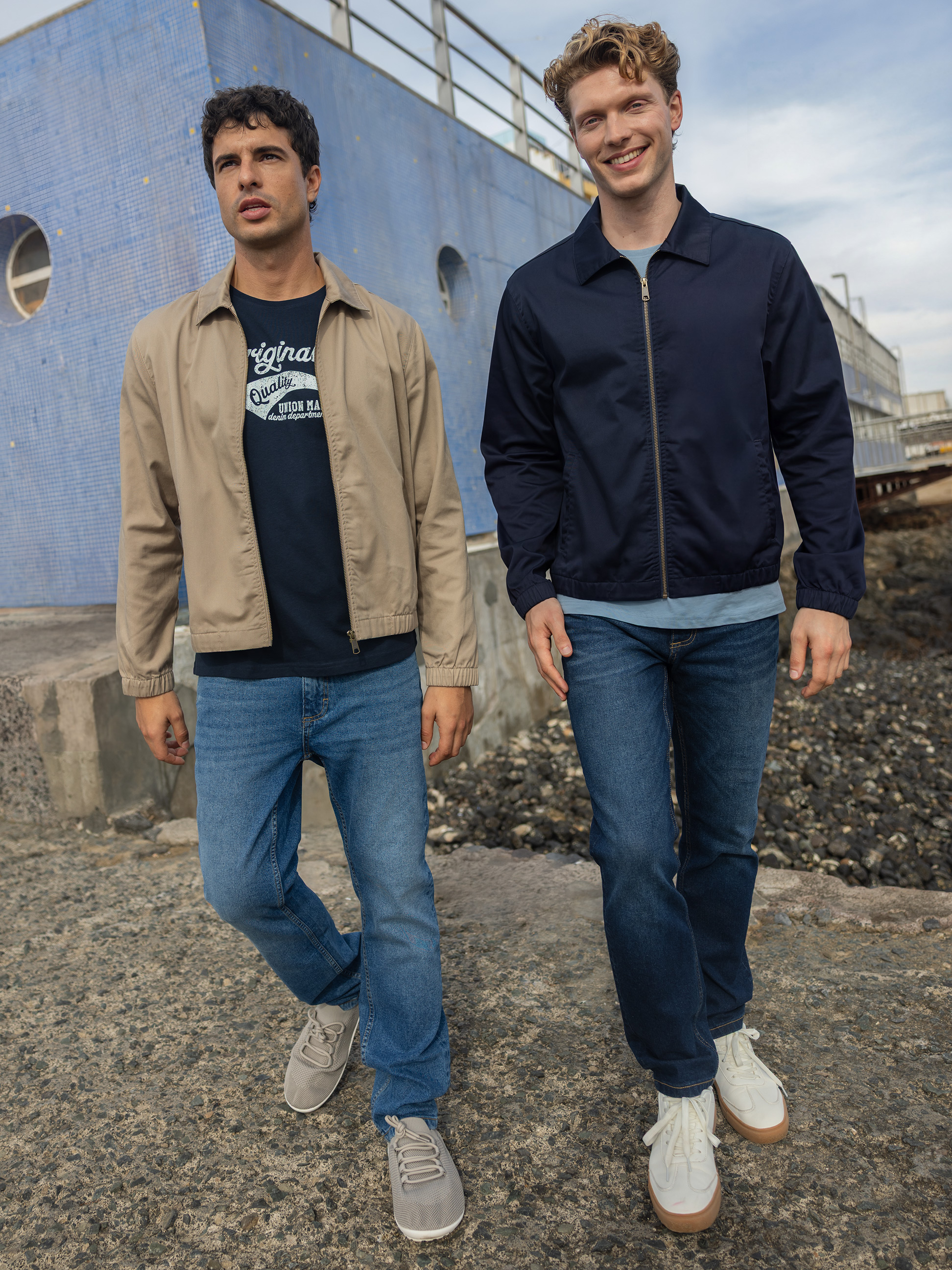 Zwei Männer in Bomberjacken und Jeans gehen an einem blauen Gebäude vorbei.