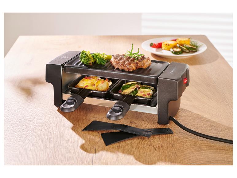 Raclette-Grill mit Steak, Brokkoli und Gemüse, plus zwei Spatel auf einem Holztisch.
