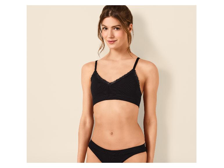 Frau in schwarzem BH und Slip mit Spitze, posiert vor einer Wand.