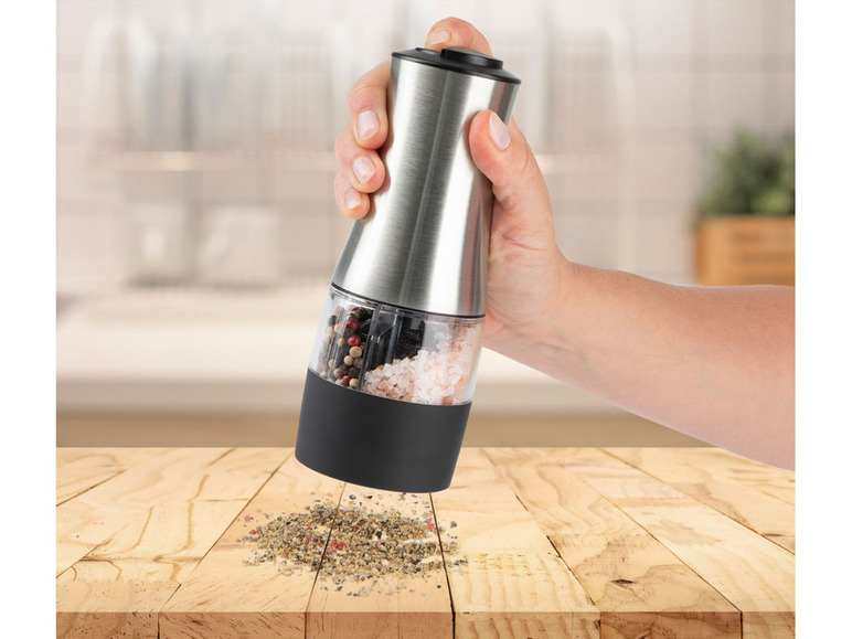 Eine Hand hält einen elektrischen Pfeffer- und Salzmühle, der gerade Salz und Pfeffer auf einen Holztisch streut.