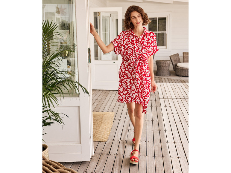 Eine Frau in einem roten Kleid mit weißem Muster geht über eine Holzterrasse.