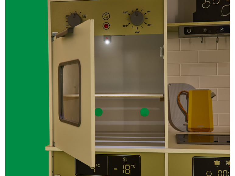 Spielküche mit offenem Backofen und Kühlschrank, mit gelber Teekanne auf der Arbeitsplatte.