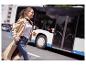 Lächelnde Frau in Trenchcoat und Jeans, mit Reisebecher, rennt an einem Bus vorbei.