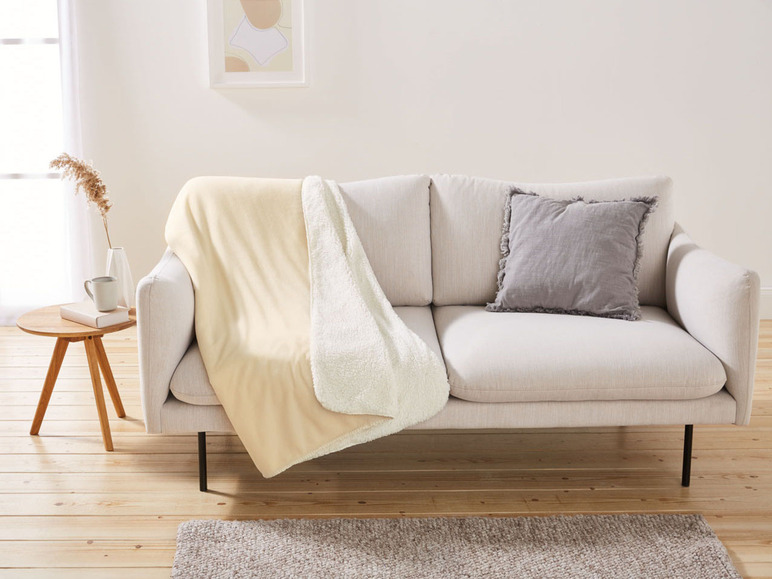 Eine beige Fleece-Decke auf einem weißen Sofa mit einem grauen Kissen.