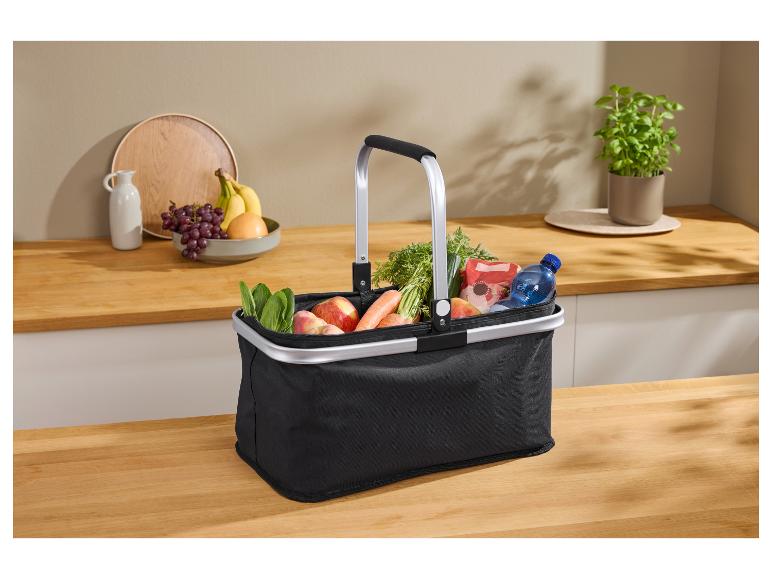 Schwarzer Einkaufskorb mit Lebensmitteln, darunter Obst, Gemüse und Wasser, auf einer Küchenarbeitsplatte.