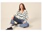 Lächelnde Frau in gestreiftem Pullover, Jeans und Loafern, auf dem Boden sitzend.