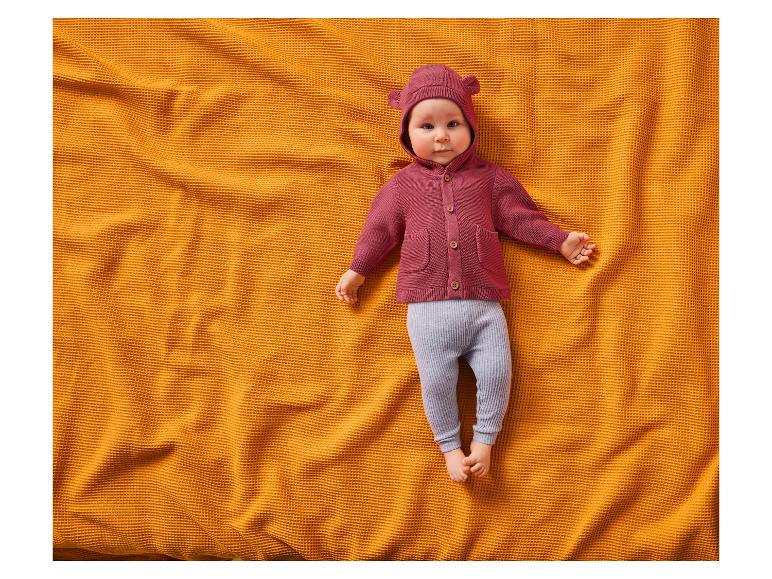 Baby in rotem Cardigan und grauer Hose auf einer orangefarbenen Waffeldecke.