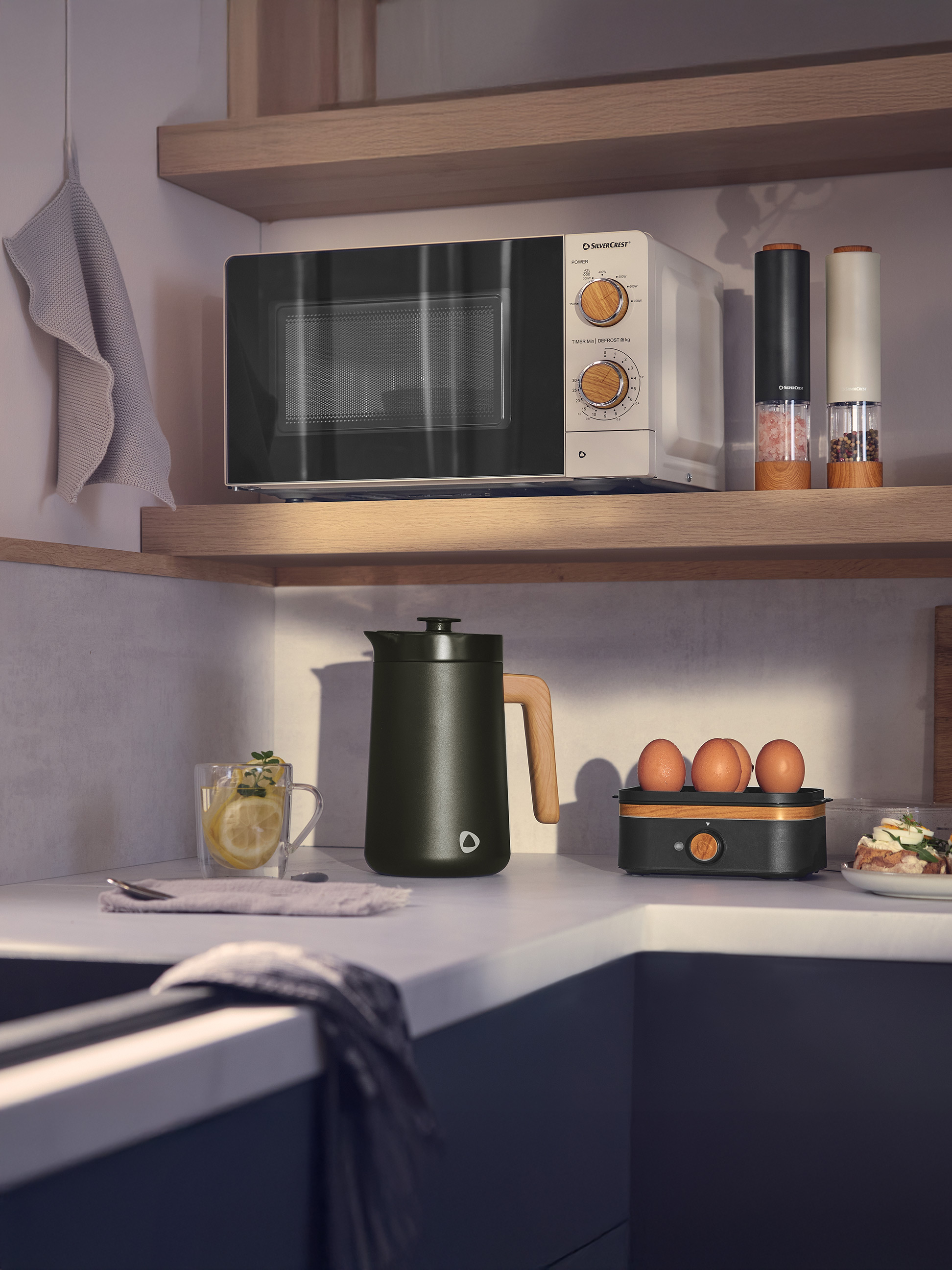 Weiß-schwarze Mikrowelle mit Holzdetails, dunkelgrüner Wasserkocher und Küchenutensilien in moderner Küche.
