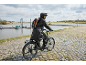 Radfahrer in schwarzem Regenmantel mit Rucksack auf einem E-Bike auf einem gepflasterten Weg.