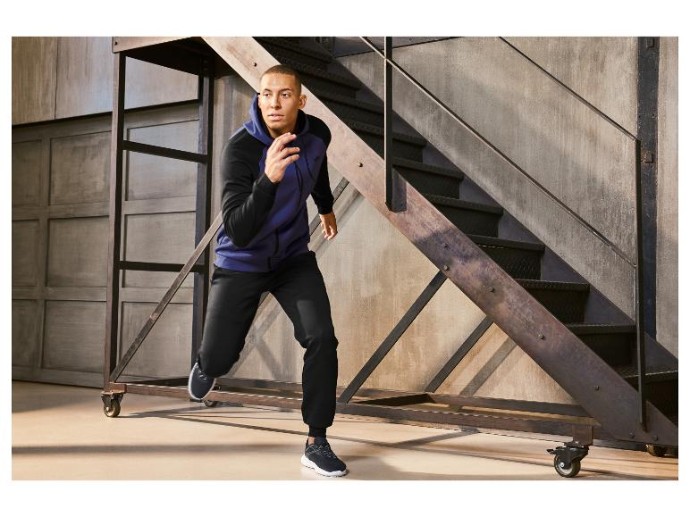 Ein Mann trägt einen blauen Kapuzenpullover und schwarze Hosen, während er auf einer Metalltreppe steht.