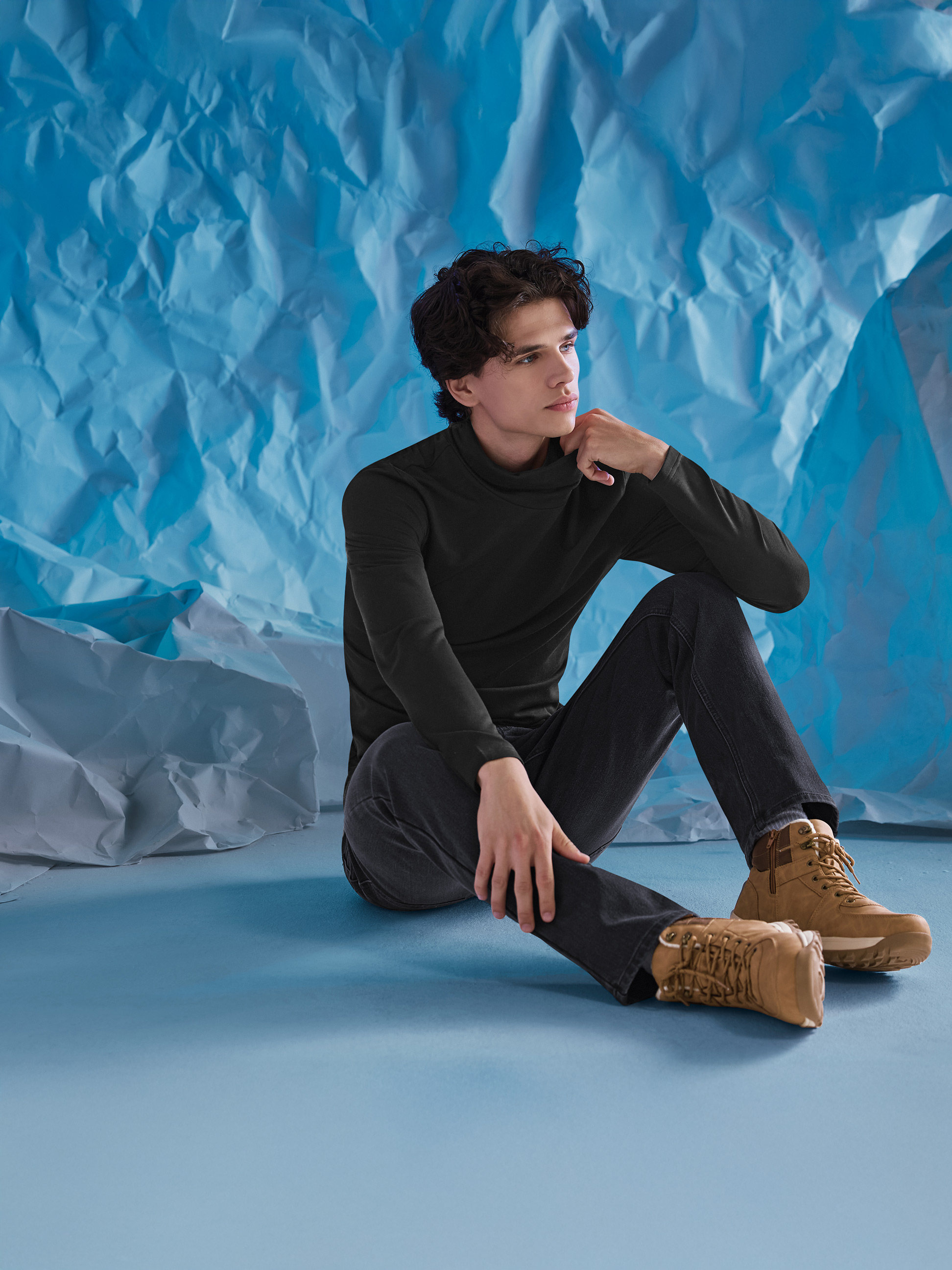 Mann in schwarzem Rollkragenpullover, Jeans und braunen Stiefeln, sitzt vor blauem Hintergrund.