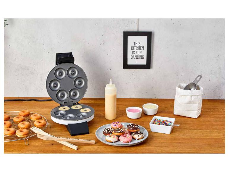 Donut-Maker mit frischen Donuts, Glasuren und Streuseln auf einem Holztisch.
