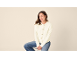 Frau in cremefarbener Strickjacke und Jeans sitzt auf einem Hocker, blickt nach rechts.