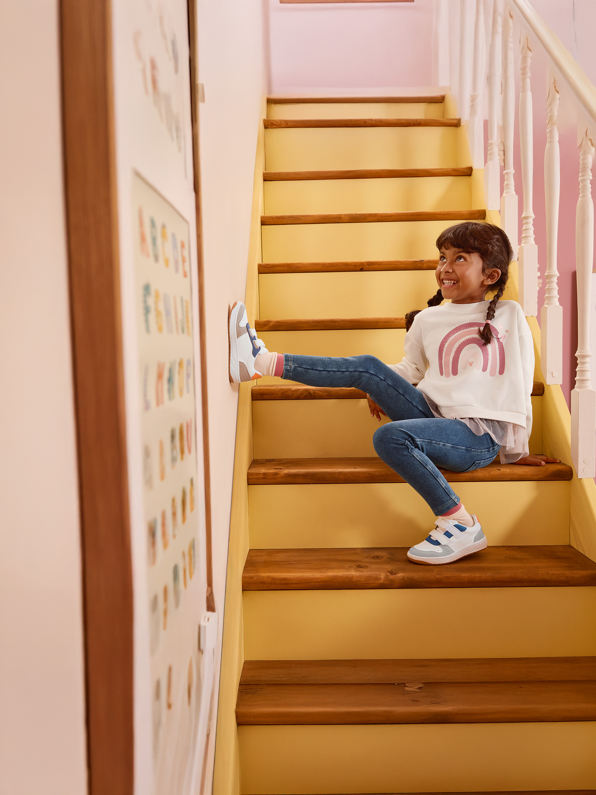 Mädchen in weißem Regenbogen-Sweatshirt und blauen Jeans sitzt auf Holztreppe