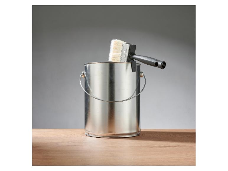 Eine metallische Farbkanne mit einem Pinsel mit schwarzem Griff darauf, auf einem Holztisch.