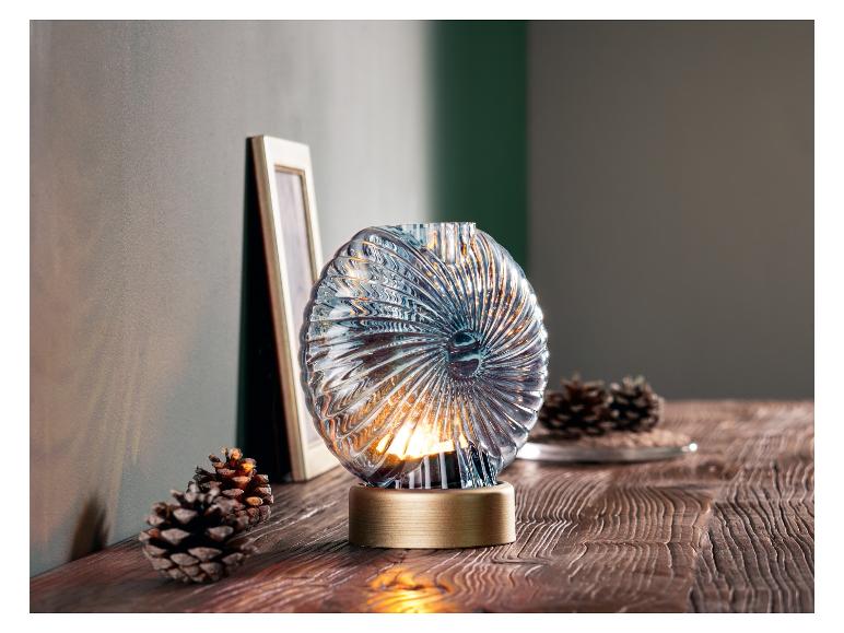 Eine gläserne Laterne mit goldenem Fuß auf einem Holztisch.