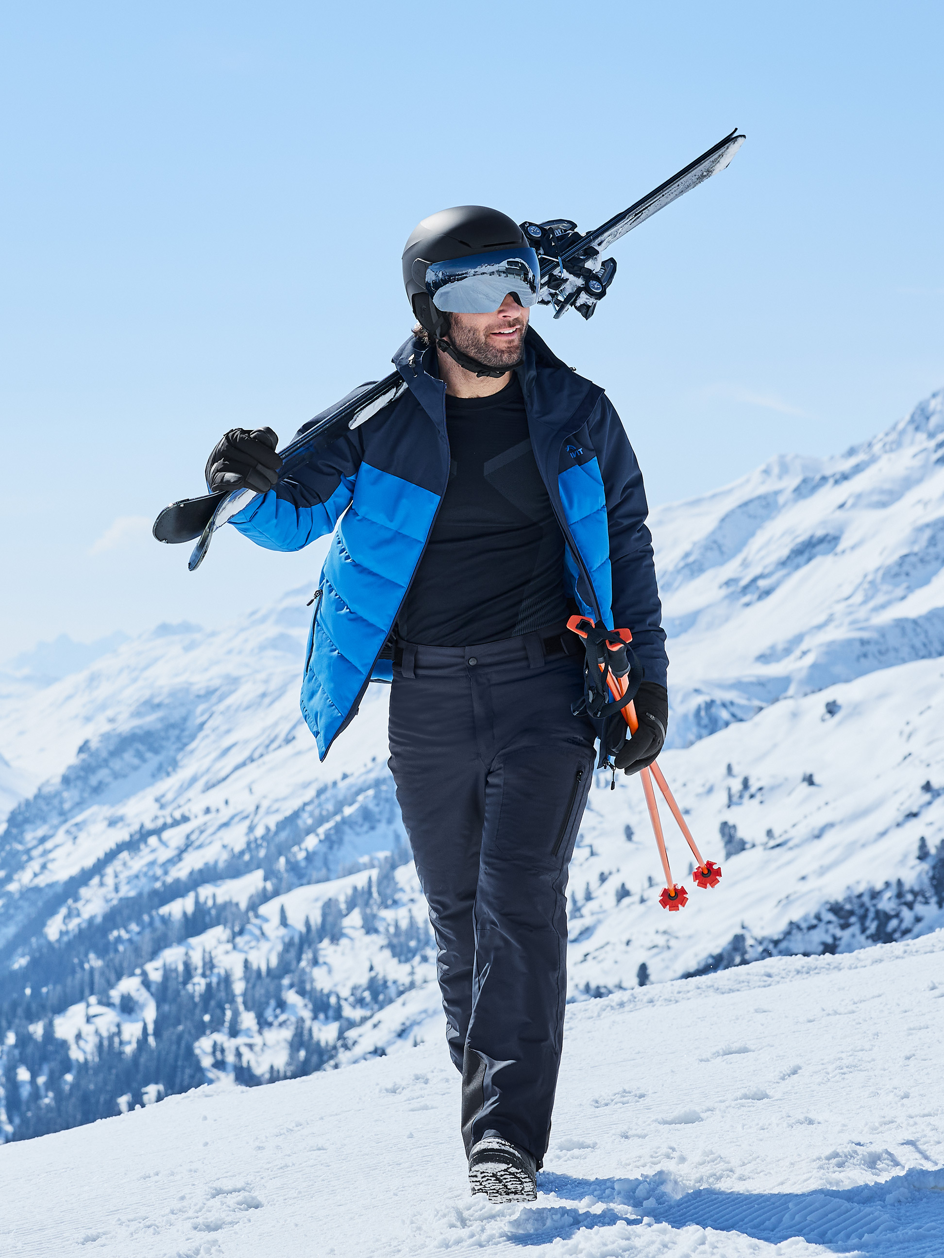 Mann in Skijacke, Skihose und Helm mit Skiern und Stöcken im Schnee.