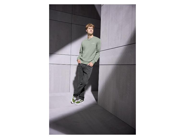 Mann in grünem Sweatshirt mit „Club 1990“ und dunkler Jeans, an eine Wand gelehnt.