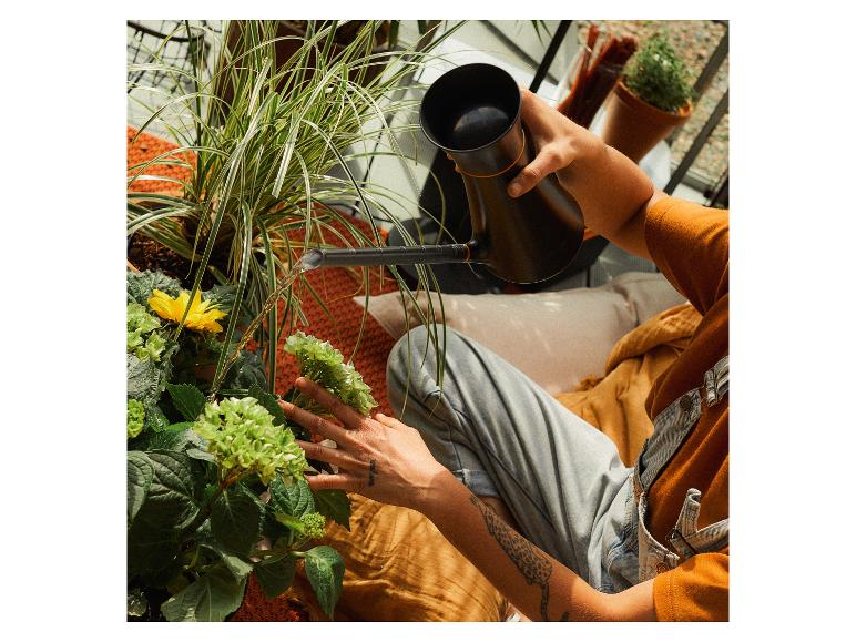 Person gießt Pflanzen auf einem Balkon, darunter eine gelbe Blume und grüne Hortensien.