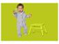 Baby im grauen Strampler mit grünem Spielzeug und gelbem Hocker auf grünem Hintergrund.