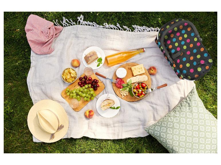 Picknickdecke mit Essen, Getränken, Hut und gepunkteter Kühltasche auf Gras.