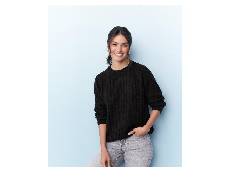 Eine Frau in einem schwarzen Pullover und grauen Jeans posiert vor einem blauen Hintergrund.