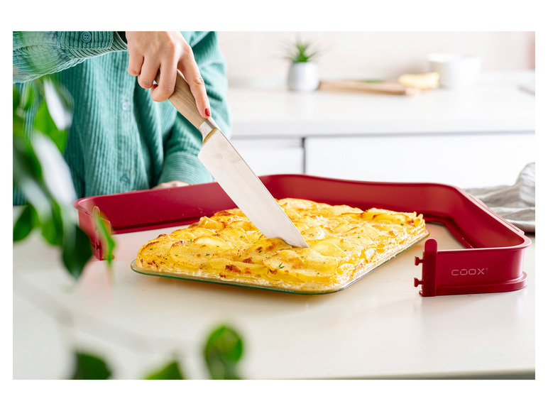 Ein gebackenes Kartoffelgericht, das mit einem Messer geschnitten wird, in einer roten Backform von Coox.