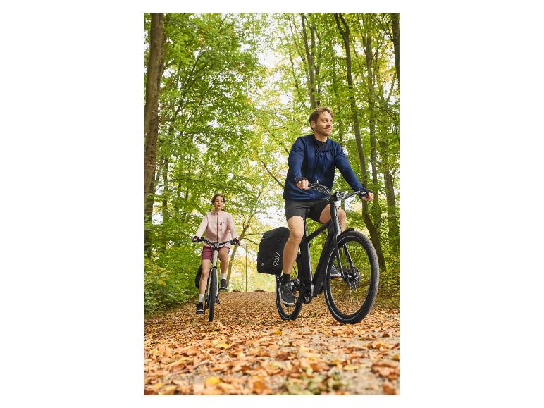 Zwei Personen radeln auf einem Waldweg, der mit Herbstlaub bedeckt ist.