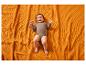 Lächelndes Baby in einem braunen Body, liegend auf einer orangefarbenen strukturierten Decke.