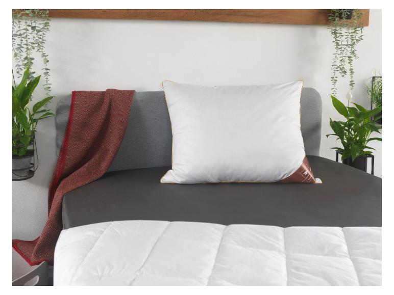 Weißes Kissen mit goldenen Rändern auf einem grauen Bett, mit Pflanzen und einer roten Decke.