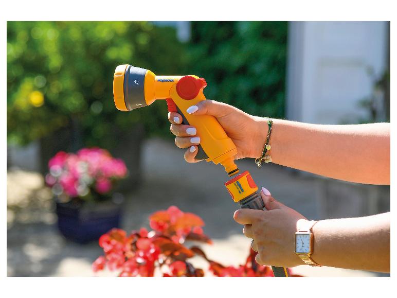 Frau hält eine gelbe Gartenspritze und verbindet sie mit einem Schlauch in einem grünen Garten.