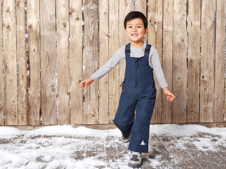 Ein Junge in einer blauen Latzhose und einem grauen Pullover steht im Schnee vor einer Holzwand.