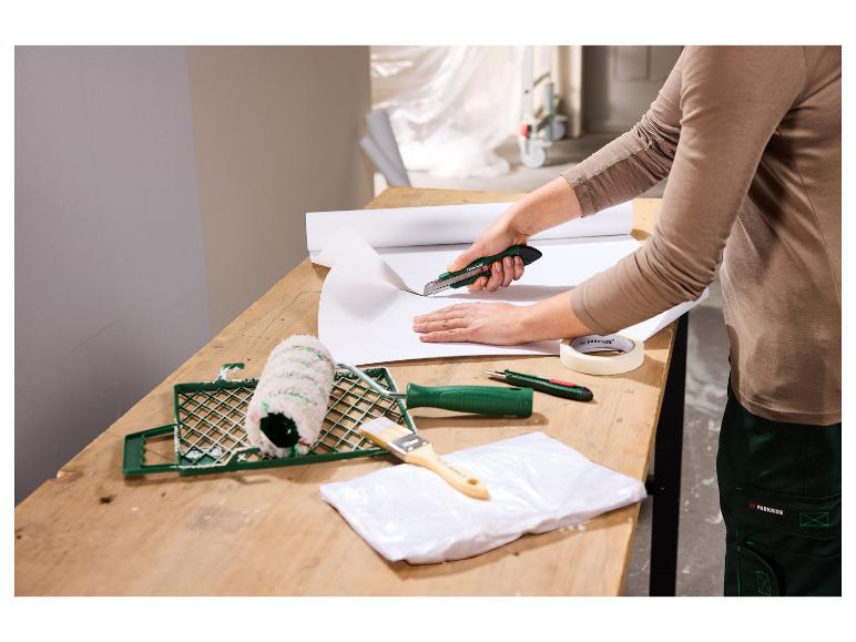 Frau schneidet Tapete mit einem Cuttermesser, mit Malerwerkzeugen auf dem Tisch.