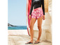 Frau in rosa Shorts mit Blumenmuster von Mistral, neben einem Surfbrett.