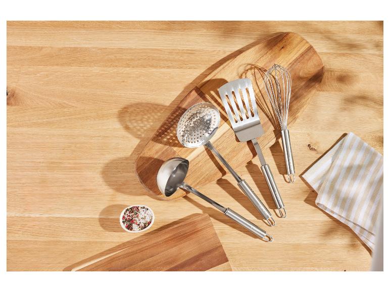 Küchenutensilien aus Edelstahl, darunter ein Pfannenwender, eine Schöpfkelle und ein Schneebesen, auf einem Holzschneidebrett.