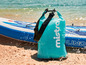 Eine türkisfarbene Drybag von Mistral mit einem aufblasbaren Paddleboard am Strand.