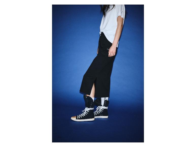 Frau in weißem T-Shirt, schwarzer Hose und High-Top-Sneakern vor blauem Hintergrund.