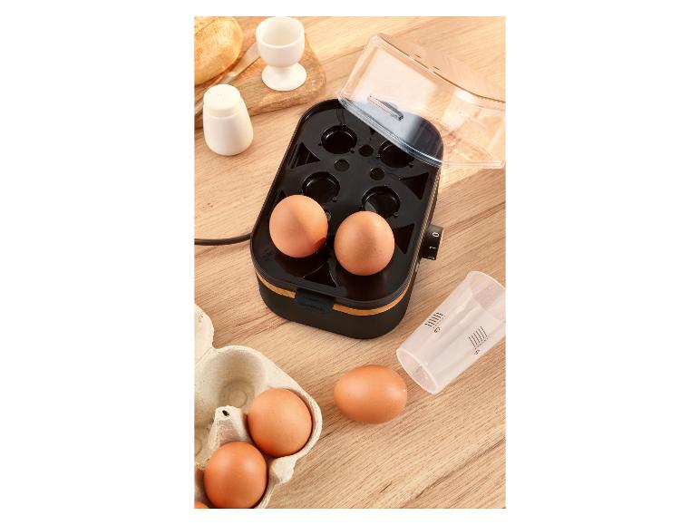 Eierkocher mit Eiern, Messbecher und Brot auf einem Holztisch.