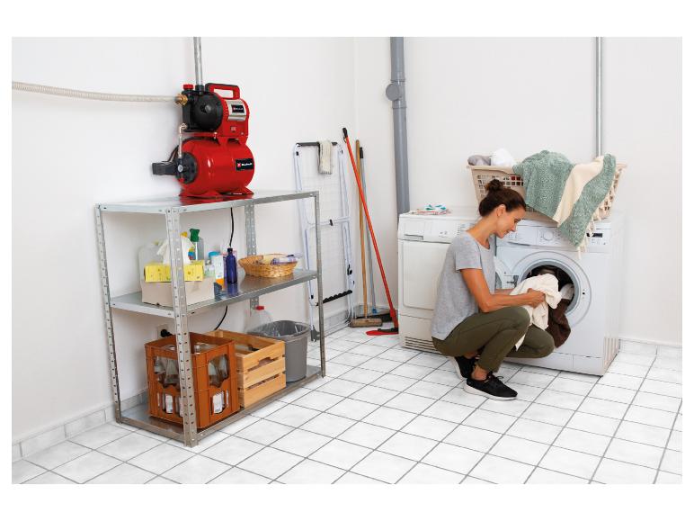 Frau belädt eine Waschmaschine in einem Hauswirtschaftsraum mit Wasserpumpe und Regalen.