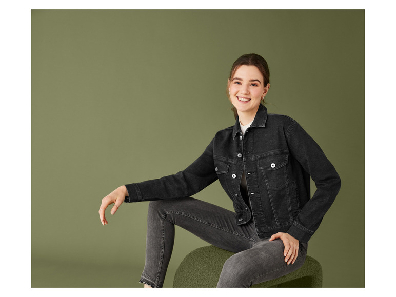 Eine Frau in einer schwarzen Jeansjacke und Jeans posiert vor einem grünen Hintergrund.