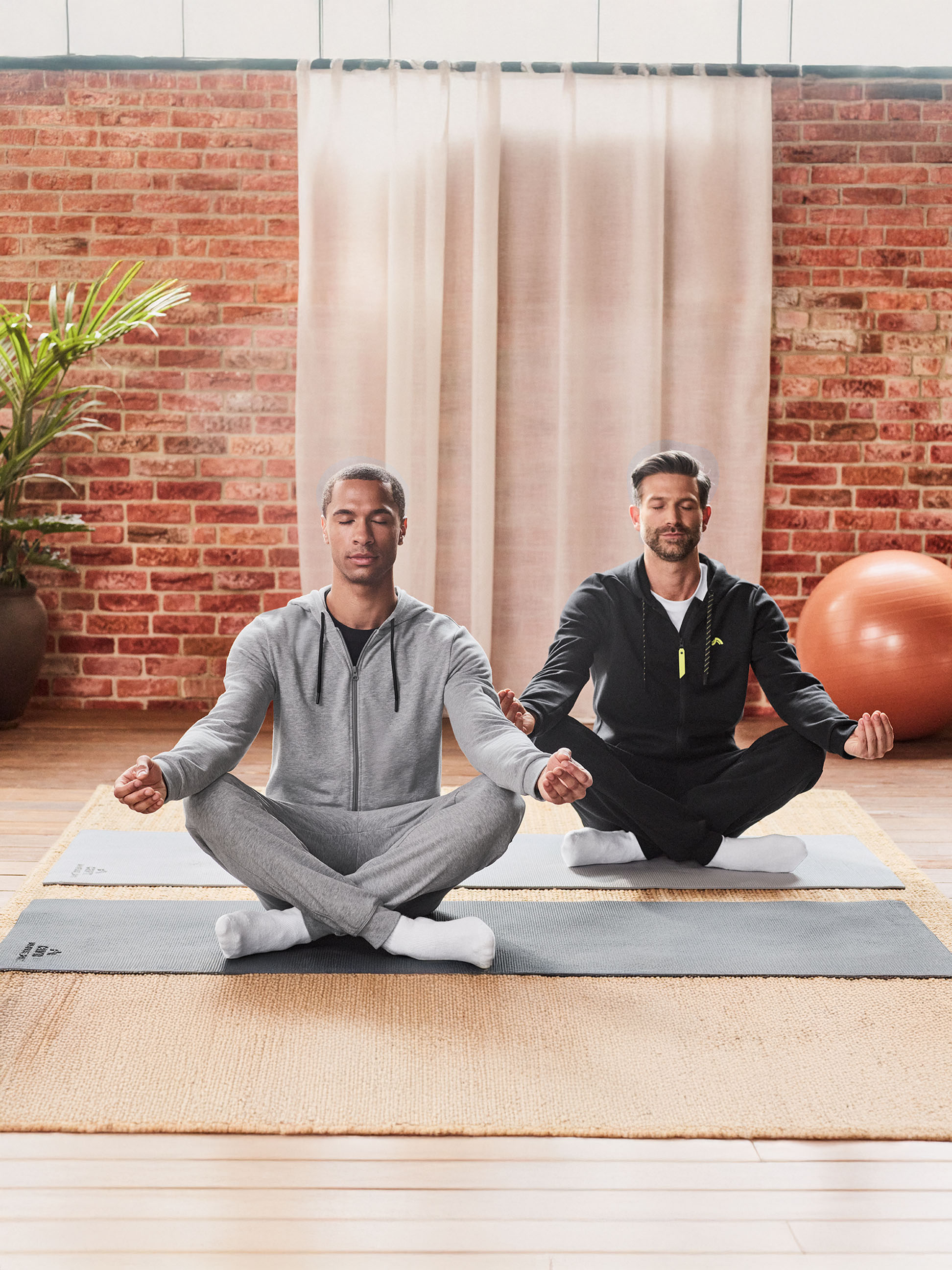Zwei Männer in Trainingsanzügen meditieren auf Yogamatten in einem Raum mit Ziegelwand.