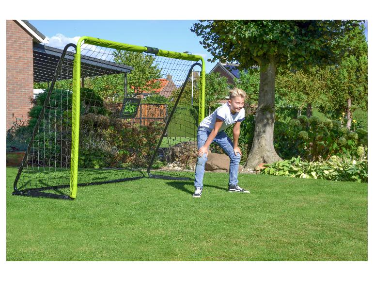 Junger Junge in Jeans und weißem T-Shirt vor einem grünen Fußballtor im Garten.