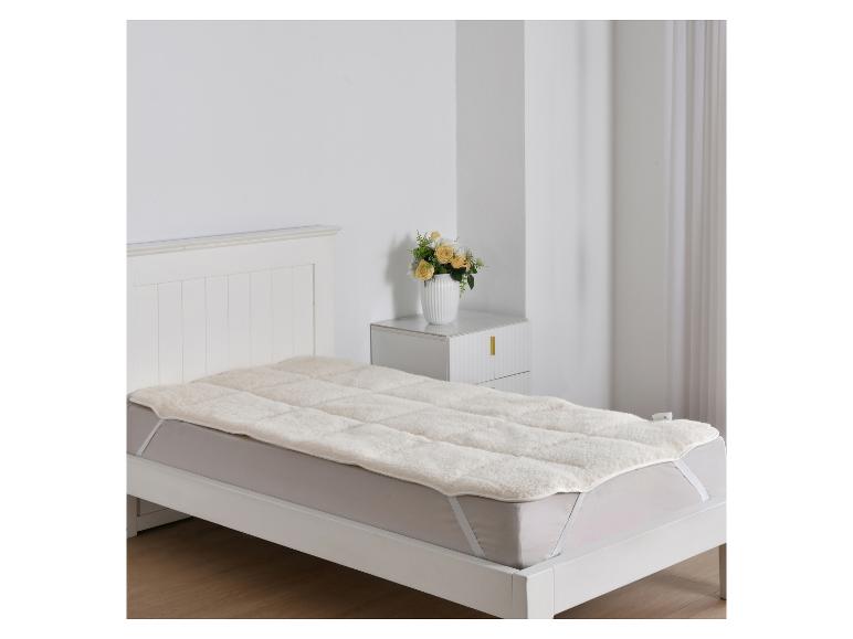 Weißes Bett mit Lammfell-Matratzenauflage und Nachttisch mit Blumen.