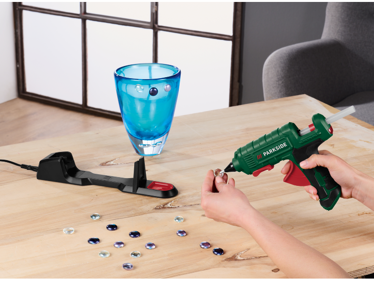 Heißklebepistole verziert eine Glasvase mit Schmucksteinen auf einem Holztisch.