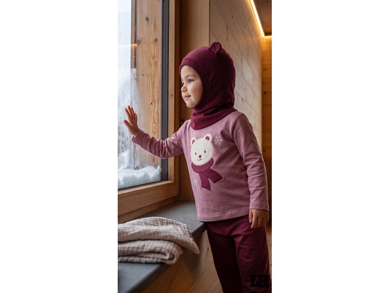Kleinkind in burgunderroter Sturmhaube und rosa Oberteil mit Eisbär-Design, schaut aus dem Fenster.