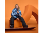 Eine Frau in blauer Skijacke und schwarzer Hose sitzt mit einem Snowboard vor orangefarbenem Hintergrund.