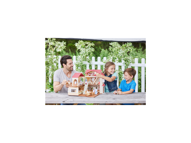Vater und zwei Kinder spielen mit einem großen Puppenhaus auf einem Holztisch im Freien.