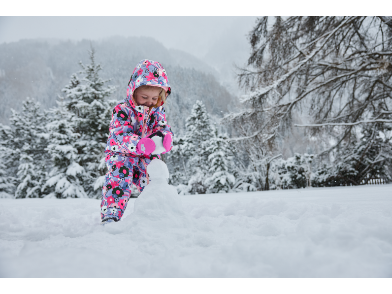 Kleinkind in buntem Schneeanzug spielt im Schnee und baut einen Schneemann.